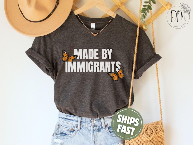 Pu&ograve; includere: Una t-shirt a maniche corte color grigio antracite scuro con la scritta bianca MADE BY IMMIGRANTS stampata al centro. Due farfalle monarca arancioni e nere sono posizionate a sinistra e a destra del testo, aggiungendo un dettaglio colorato.