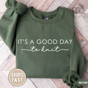May include: Military green sweatshirt with the text "IT'S A GOOD DAY to knit" in white script. The long-sleeved crewneck top has a round, ribbed collar and cuffs. A round sticker with the words "SHIPS FAST" is on the bottom left.