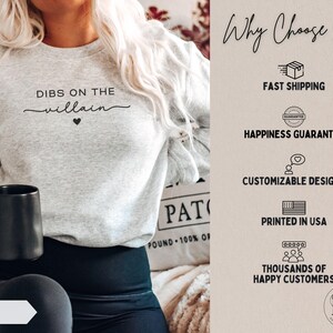 May include: Light gray sweatshirt with the text "DIBS ON THE villain" in black script and a small black heart. The word "ASH" is visible on a white arrow. A black mug is held in the hand.