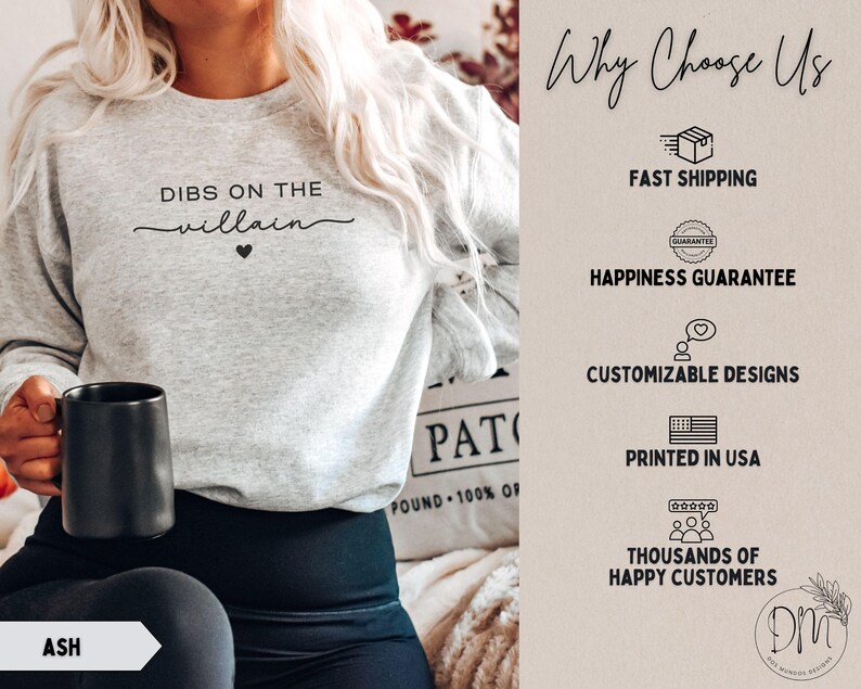 May include: Light gray sweatshirt with the text "DIBS ON THE villain" in black script and a small black heart. The word "ASH" is visible on a white arrow. A black mug is held in the hand.