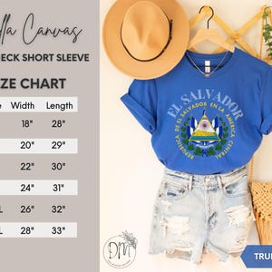 May include: A true royal blue short-sleeve crewneck t-shirt featuring the El Salvador coat of arms graphic. The image includes a size chart with width and length measurements in inches for sizes S through 3XL, a tan hat, and denim shorts.
