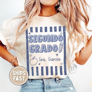 May include: An ivory t-shirt featuring a blue graphic design with vertical stripes, the text "SEGUNDO GRADO", a pencil icon, an apple icon, and the text "Sra. Garcia". The design includes small star accents and is centered on the front of the shirt.