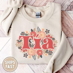 May include: Sand-colored sweatshirt with the word "T&iacute;a" in red, surrounded by a floral design in shades of pink, peach, and navy. The sweatshirt is made of a soft material and has a crew neck.