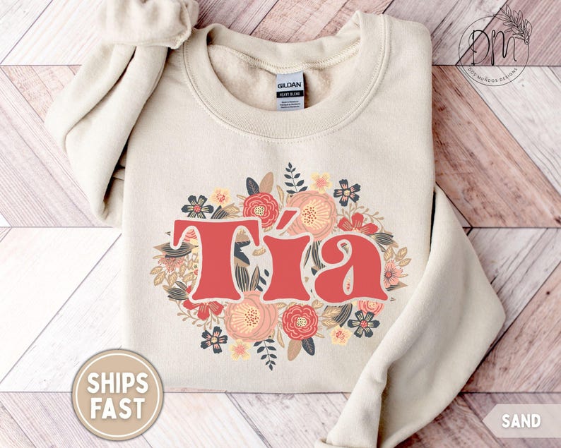 May include: Sand-colored sweatshirt with the word "T&iacute;a" in red, surrounded by a floral design in shades of pink, peach, and navy. The sweatshirt is made of a soft material and has a crew neck.