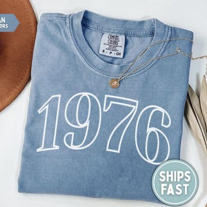 May include: A blue jean Comfort Colors t-shirt with the year "1976" printed in white. A gold necklace with a pendant rests on the shirt. A brown hat and dried plant stems are in the background.