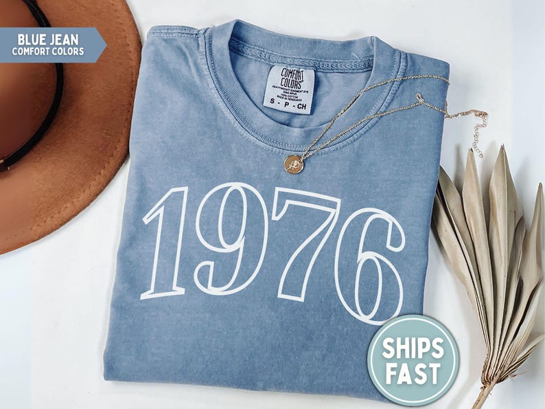 May include: A blue jean Comfort Colors t-shirt with the year "1976" printed in white. A gold necklace with a pendant rests on the shirt. A brown hat and dried plant stems are in the background.
