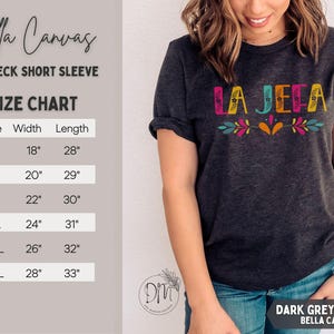 May include: Dark grey heather Bella Canvas crewneck short sleeve t-shirt with the text "LA JEFA" in colorful letters. The size chart shows measurements in inches: S (45.7cm width, 71.1cm length), M (50.8cm width, 73.7cm length), L (55.9cm width, 76.2cm length), XL (61cm width, 78.7cm length), 2XL (66cm width, 81.3cm length), 3XL (71.1cm width, 83.8cm length).