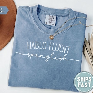 K&ouml;nnte beinhalten: Blaues Jeans-T-Shirt mit dem wei&szlig;en Schriftzug "HABLO FLUENT spanglish". Eine goldene Halskette mit Anh&auml;nger liegt auf dem Shirt. Ein brauner Hut und getrocknete Pflanzen sind im Hintergrund.