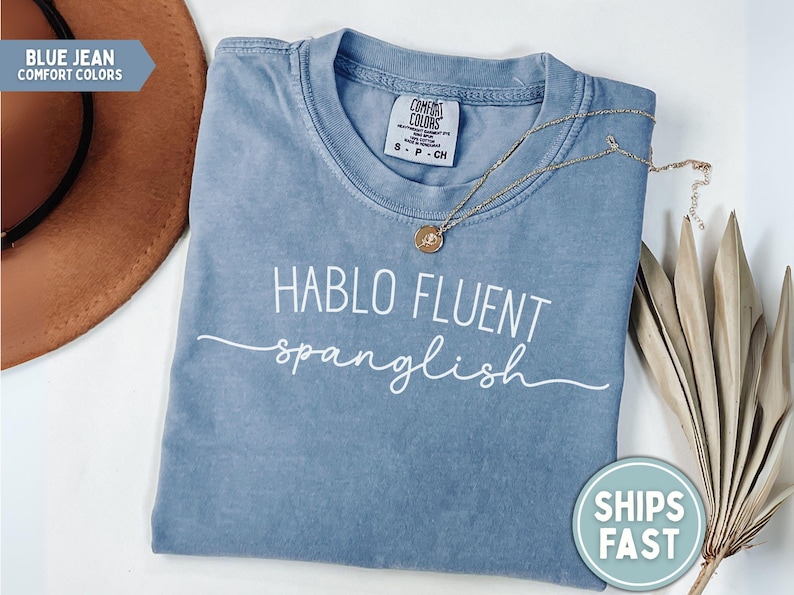 K&ouml;nnte beinhalten: Blaues Jeans-T-Shirt mit dem wei&szlig;en Schriftzug "HABLO FLUENT spanglish". Eine goldene Halskette mit Anh&auml;nger liegt auf dem Shirt. Ein brauner Hut und getrocknete Pflanzen sind im Hintergrund.
