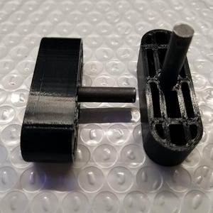 May include: Two black, 3D-printed components with cylindrical rods. One component is rectangular with a rod extending through it. The other is a similar shape, but with a hollowed-out design and a rod inserted.