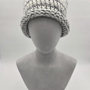 May include: A thick-knit beanie hat in a speckled pattern of white, grey, and black. The hat has a folded brim and a rounded crown. The hat is displayed on a grey mannequin head against a white background.