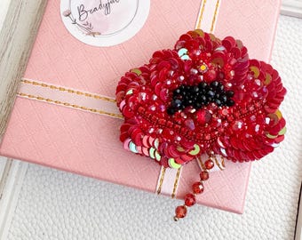 Könnte beinhalten: Rote, paillettenbesetzte Mohnblumenbrosche mit schwarzen Perlenakzenten, die auf einer rosa Geschenkbox liegt. Die Brosche hat einen Perlenstiel und ist mit schillernden Pailletten verziert. Die Box ist mit einem goldenen Band und einem runden Aufkleber mit dem Text "Beadyjulia" verziert.