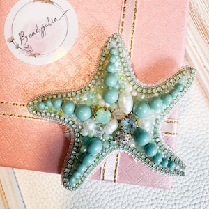 Puede incluir: Broche de estrella de mar con cuentas en tonos turquesa, verde mar y blanco, con detalles de perlas y cristales. El broche está colocado sobre una caja de regalo rosa con una cinta dorada. Hecho a mano.
