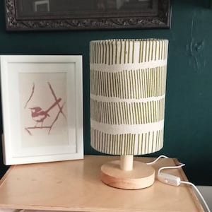 May include: A white table lamp with a green and white patterned fabric shade. The shade has a geometric pattern of vertical lines. The lamp is on a wooden table next to a white picture frame with a bird illustration.