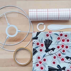 Op de afbeelding: Een set benodigdheden voor het maken van een lampenkap. De benodigdheden omvatten een wit lampenkapframe, een rol wit gekarteld papier, een rol bruin tape, een witte kunststof schraper, een witte kunststof ring en een stuk stof met een blauwe achtergrond en een patroon van zwarte vogels en rode bloemen.