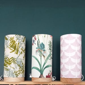 Op de afbeelding: Drie cilindrische tafellampen met verschillende bloemenpatronen. De lampen hebben witte bases met een houtlook. De eerste lamp heeft een groen en wit bloemenpatroon, de tweede lamp heeft een groen en wit bloemenpatroon met een blauwe bloem en de derde lamp heeft een roze en wit bloemenpatroon.