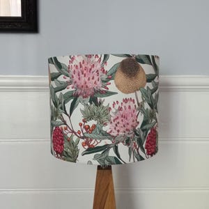 May include: A white lampshade with a floral print featuring pink and red flowers, green leaves, and brown seed pods. The lampshade is on a wooden lamp base.