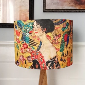 May include: A lampshade with a portrait of a woman holding a fan, set against a backdrop of floral and peacock motifs. The design incorporates gold, red, blue, and green, creating an artistic element for home decor.