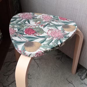 May include: A small, wooden stool with a floral patterned top. The top features pink flowers, green leaves, and red berries on a white background. The legs are a light wood color, and the stool is near a gray couch.