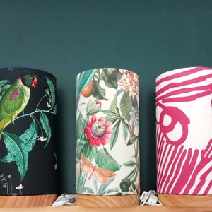 Könnte beinhalten: Drei zylindrische Tischlampen mit verschiedenen gemusterten Stoffschirmen. Die Lampenschirme zeigen einen Papagei auf einem Ast, tropische Blumen und abstrakte rosa Linien.