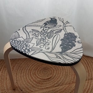 May include: A wooden stool with a triangular seat featuring a black and white floral pattern with a bird design. The stool is sitting on a woven rug.