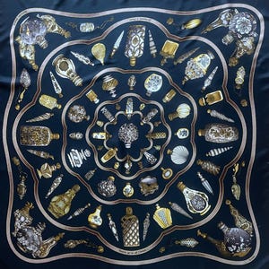 May include: A square, navy blue silk scarf featuring a repeating pattern of ornate perfume bottles in gold, white, and silver. The bottles vary in shape and design, with intricate detailing. A brown border frames the central design.