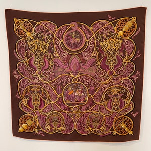 May include: A brown silk scarf with an intricate gold and purple design. The design features a repeating pattern of stylised flowers, leaves, and birds. The scarf is bordered with a gold trim.