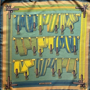 Puede incluir: Pañuelo de seda con un patrón repetido de cintas coloridas en tonos verdes, amarillos y azules. Las cintas están dispuestas sobre barras doradas sobre un fondo turquesa, enmarcadas por un borde beige y verde azulado oscuro. El texto "HERMES-PARIS" es visible.