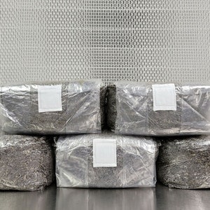 May include: Five rectangular blocks of compressed, dark gray material wrapped in clear plastic with white labels. The blocks are stacked in a pyramid shape on a silver surface.
