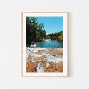 Może przedstawiać: Zdjęcie w ramie przedstawiające mężczyznę skaczącego do błękitnej rzeki z kamiennym wodospadem na pierwszym planie. Rzeka otoczona jest zielonymi drzewami i błękitnym niebem.