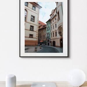K&ouml;nnte beinhalten: Ein gerahmtes Foto einer engen Stra&szlig;e in Prag, Tschechien. Die Stra&szlig;e ist ges&auml;umt von bunten Geb&auml;uden, einige mit gew&ouml;lbten Durchg&auml;ngen. Die Geb&auml;ude sind in Gelb-, Gr&uuml;n- und Rosat&ouml;nen gestrichen. Die Stra&szlig;e ist mit Kopfsteinpflaster ausgelegt und es sind ein paar Leute auf der Stra&szlig;e unterwegs.