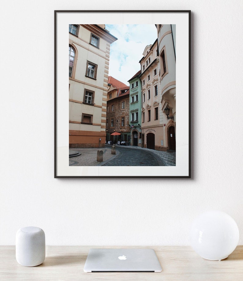 K&ouml;nnte beinhalten: Ein gerahmtes Foto einer engen Stra&szlig;e in Prag, Tschechien. Die Stra&szlig;e ist ges&auml;umt von bunten Geb&auml;uden, einige mit gew&ouml;lbten Durchg&auml;ngen. Die Geb&auml;ude sind in Gelb-, Gr&uuml;n- und Rosat&ouml;nen gestrichen. Die Stra&szlig;e ist mit Kopfsteinpflaster ausgelegt und es sind ein paar Leute auf der Stra&szlig;e unterwegs.