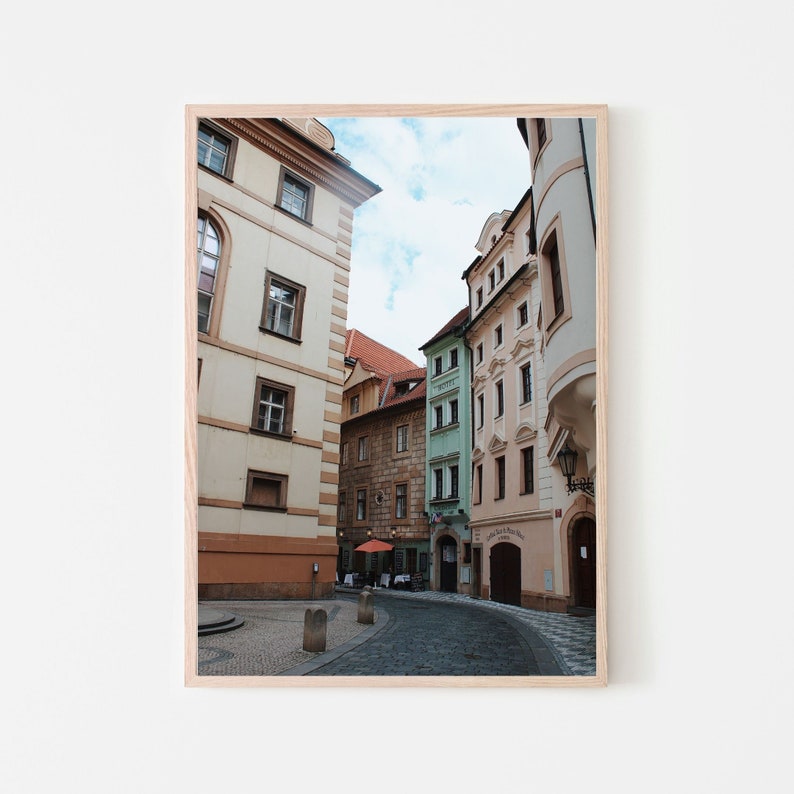 K&ouml;nnte beinhalten: Eine schmale Kopfsteinpflasterstra&szlig;e in Prag, Tschechien, ges&auml;umt von bunten Geb&auml;uden. Die Geb&auml;ude sind eine Mischung aus Gelb, Rosa und Gr&uuml;n, und einige haben verzierte Details. Die Stra&szlig;e ist leer, bis auf ein paar Tische und St&uuml;hle, die vor einem Caf&eacute; aufgestellt sind.