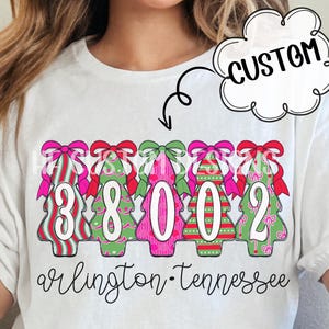 May include: White t-shirt featuring a festive graphic of Christmas tree-shaped numbers and bows. The numbers are 38002, with the words "arlington" and "tennessee" printed below. A "CUSTOM" speech bubble is in the upper right.