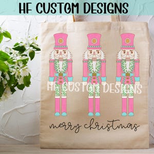 May include: A pink tote bag with three pink nutcracker figures with green and white accents. The bag has the text "HF Custom Designs" and "Merry Christmas" printed on it.