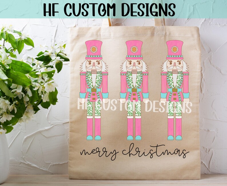May include: A pink tote bag with three pink nutcracker figures with green and white accents. The bag has the text "HF Custom Designs" and "Merry Christmas" printed on it.