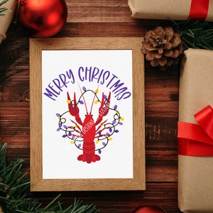 May include: A framed print with a red crawfish with purple and yellow lights wrapped around it. The text "Merry Christmas" is written in purple letters on a white background.