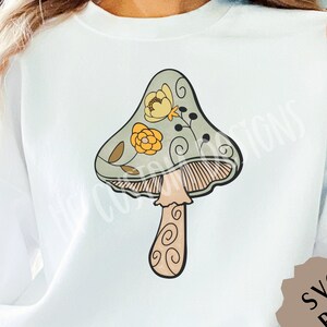 May include: A white sweatshirt with a green, yellow, and brown mushroom graphic with floral details. The mushroom has a stem with a swirl pattern.