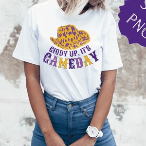 May include: White t-shirt featuring a purple and gold leopard print cowboy hat graphic with the text "Giddy Up, It's Gameday".