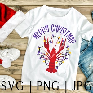 May include: White t-shirt with a red crawfish design and the text "Merry Christmas" in purple letters. The crawfish is surrounded by purple and yellow Christmas lights.