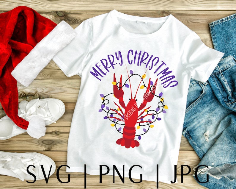 May include: White t-shirt with a red crawfish design and the text "Merry Christmas" in purple letters. The crawfish is surrounded by purple and yellow Christmas lights.