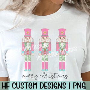 May include: A white t-shirt with a pink, green, and blue design of three nutcracker soldiers. The text "merry christmas" is printed below the nutcrackers.