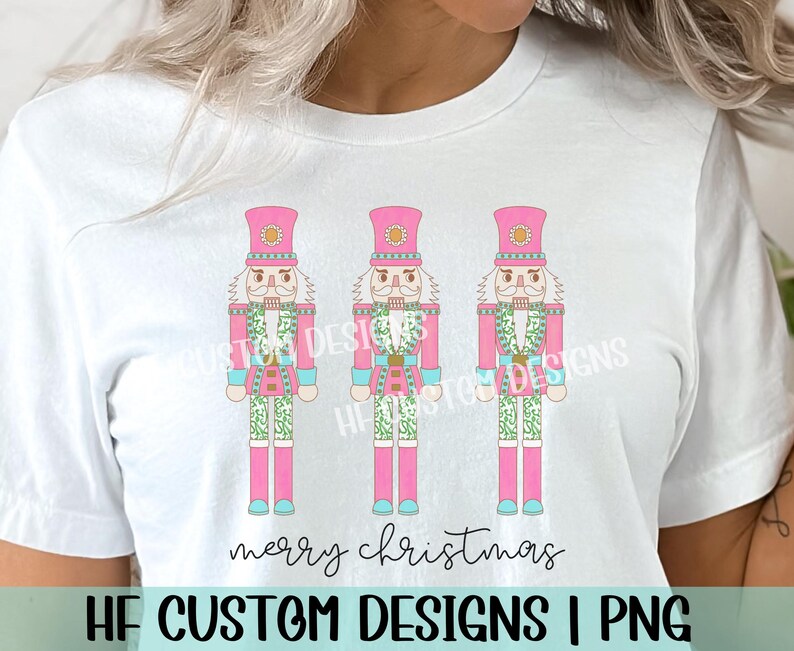 May include: A white t-shirt with a pink, green, and blue design of three nutcracker soldiers. The text "merry christmas" is printed below the nutcrackers.