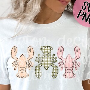 May include: A white t-shirt with three different colored lobsters on it. The lobsters are in a row, and each one is a different color: pink, green, and pink. The lobsters are all wearing a checkered pattern.