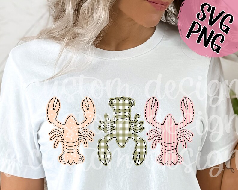 May include: A white t-shirt with three different colored lobsters on it. The lobsters are in a row, and each one is a different color: pink, green, and pink. The lobsters are all wearing a checkered pattern.