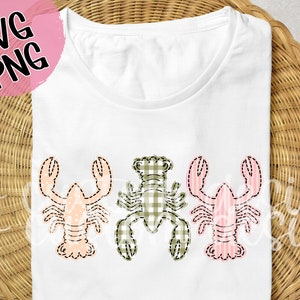 May include: A white t-shirt with three embroidered lobsters. The lobsters are in a row, with the middle lobster in green and white gingham and the two outer lobsters in pink and white stripes.