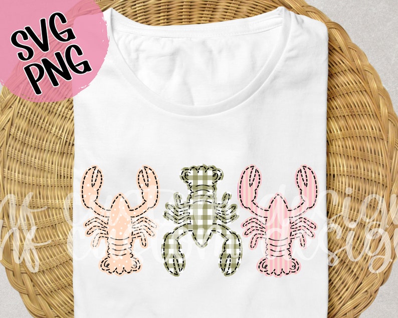 May include: A white t-shirt with three embroidered lobsters. The lobsters are in a row, with the middle lobster in green and white gingham and the two outer lobsters in pink and white stripes.