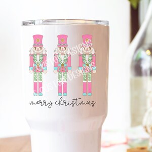 May include: White tumbler with a pink background and three nutcracker illustrations. The nutcrackers are wearing pink hats and have green and blue accents. The text "merry christmas" is written in black at the bottom of the tumbler.