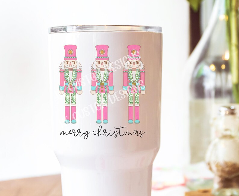 May include: White tumbler with a pink background and three nutcracker illustrations. The nutcrackers are wearing pink hats and have green and blue accents. The text "merry christmas" is written in black at the bottom of the tumbler.