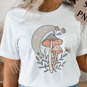 May include: White t-shirt with a graphic of two mushrooms, a crescent moon, and flowers. The mushrooms are orange and brown with white spots. The moon is gray with white stars. The flowers are pink, yellow, and white.